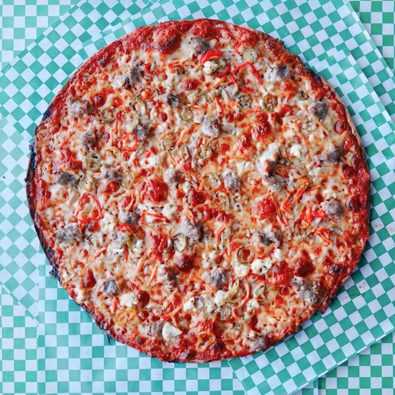 A whole pizza with sausage, peppers, and jalapeños on a green and white checkered paper.